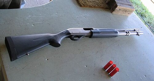 A Remington 870 pump‑action shotgun with a black synthetic stock and fore‑end resting on a shooting bench, with three red shotgun shells placed beside it.