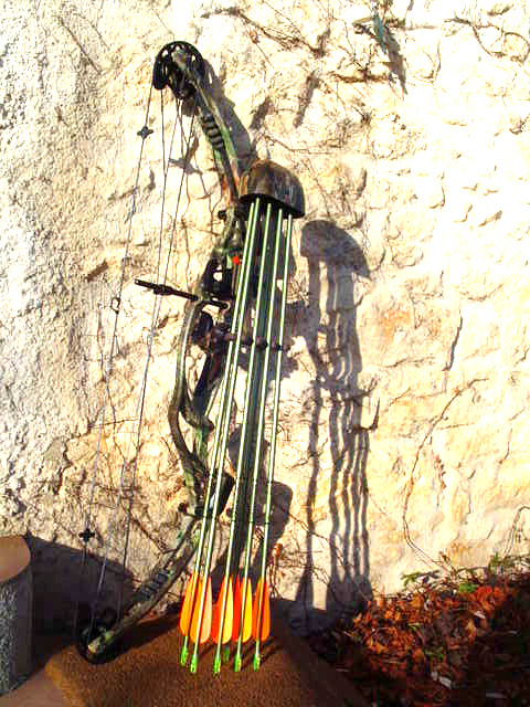 A camouflage compound bow leaning against a stone wall, equipped with a quiver holding five arrows with orange fletching and green shafts.
